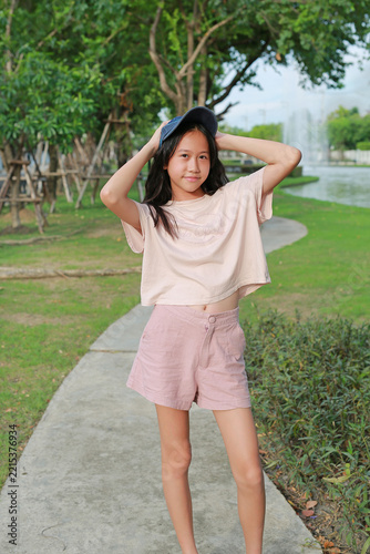 A young Asian girl wearing a casual pink top, shorts, and a cap posing on a pathway in a park.