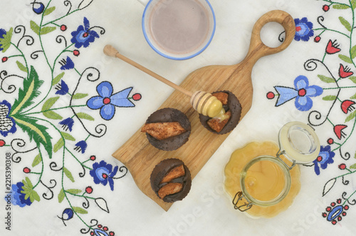 Three chocolate muffins with fruit, a honey drizzling onto cupcake, a cup of cocoa and glass jar of honey on embroidered tablecloth with floral motifs.