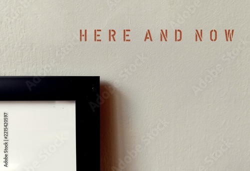 Photo Frame on a wall with text phrase HERE AND NOW - refers to present moment, being conscious and mindful of what happening right now than on past or worrying about future