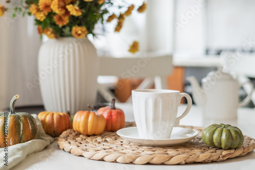White coffee cup surrounded by colorful miniature pumpkins and orange flowers on a bright kitchen table, suggesting a cozy autumn morning at home.