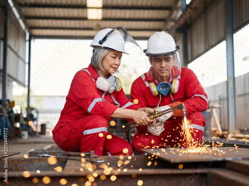 Mature female welder guiding a young apprentice with a cutting torch, illustrating industrial mentorship and vocational training.