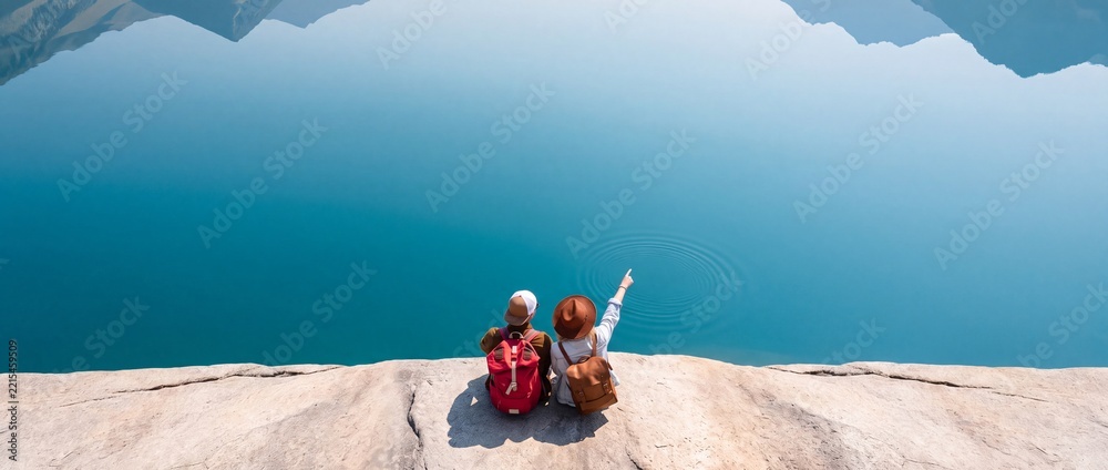 Fototapeta premium High angle rear view of a young couple with backpacks sitting on a cliff edge, pointing at a ripple on the surface of a crystal clear blue mountain lake with scenic mountain reflection.