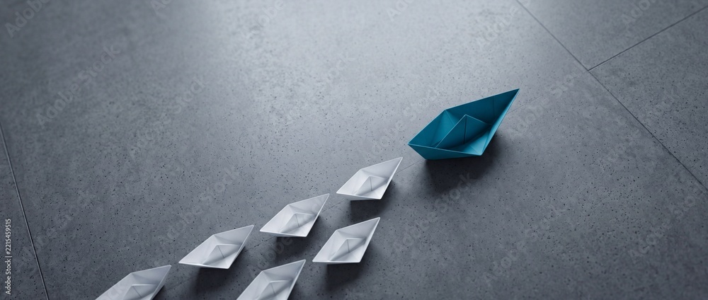 Fototapeta premium Leading the Way Concept: A Single Blue Paper Boat Navigating Forward Followed by a Group of White Paper Boats on a Gray Stone Floor, Representing Leadership, Teamwork, and Business Strategy.