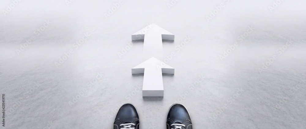 Fototapeta premium High angle point of view shot of person standing in front of two white arrows on grey floor, representing progress, direction, decision making, perspective shift, and future career movement.