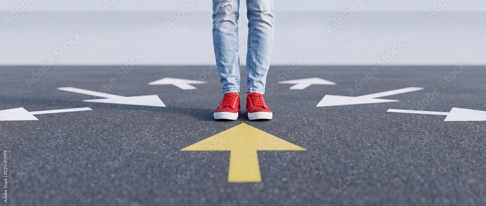 Fototapeta premium Person wearing red sneakers standing on asphalt road at a crossroads with multiple white arrows and one distinct yellow arrow pointing forward representing choice, decision, and a new perspective.