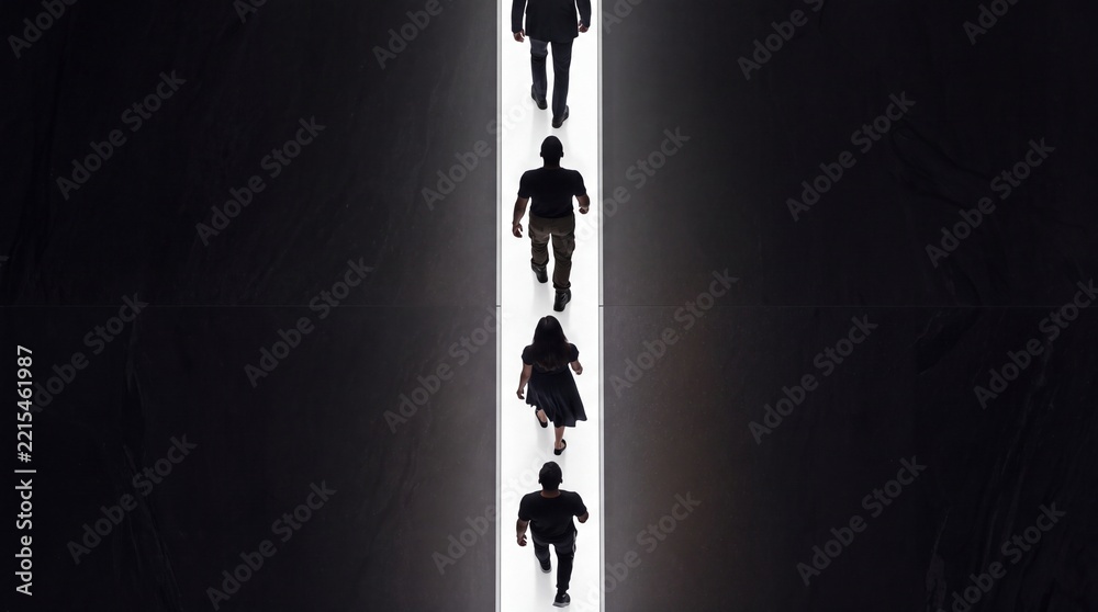 Fototapeta premium Top down overhead view of diverse people walking in a single file line along a bright glowing path in a dark minimalist space, representing direction, progress, and conceptual leadership.