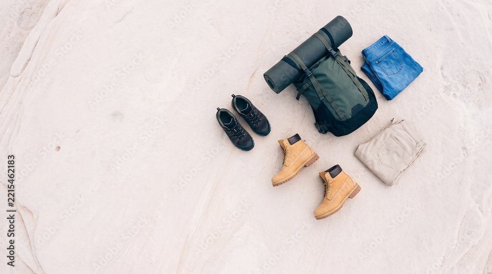 Fototapeta premium Top down overhead view of essential hiking and travel equipment including a backpack, rolled mat, leather boots, trekking shoes, and folded clothes laid out on a textured natural rock surface.
