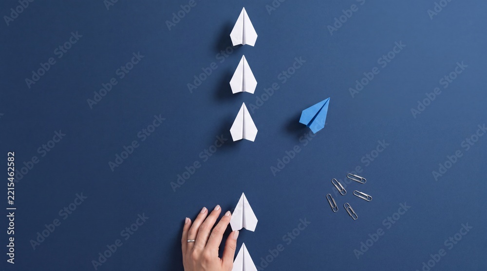 Fototapeta premium Top Down View of a Hand Placing White Paper Planes in a Row with One Blue Paper Plane Changing Direction on a Dark Blue Background, Concept of Leadership, Uniqueness, and Business Innovation