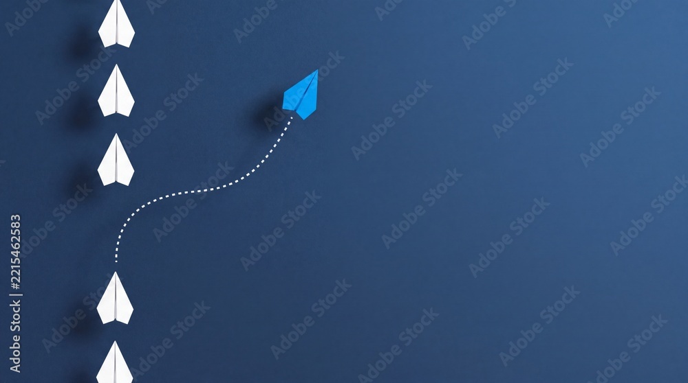 Fototapeta premium Overhead View of a Blue Paper Airplane Changing Direction and Deviating from a Group of White Airplanes on a Blue Background, Representing Unique Leadership, Innovation, and Business Strategy.