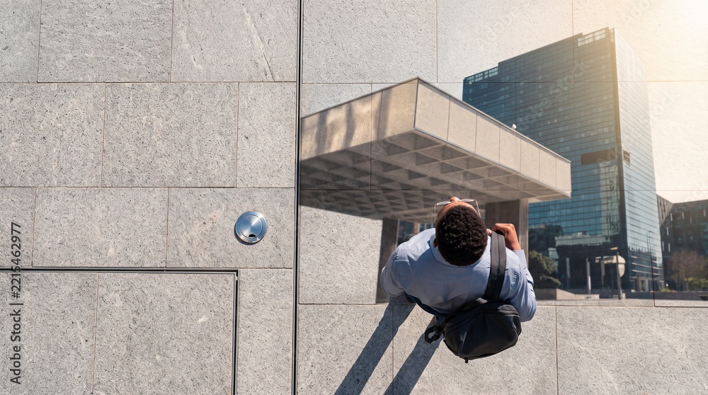 Fototapeta premium Top down view of a professional man with a backpack walking along a grey stone pavement next to a skyscraper reflection in a modern city district during a bright sunny day.