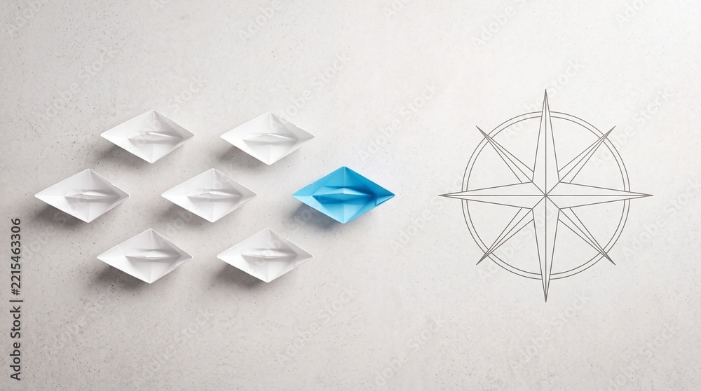 Fototapeta premium Top down overhead view of a blue paper boat leading a fleet of white boats towards a compass symbol, representing leadership, business strategy, guidance, and unique direction on a grey background.