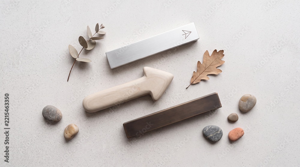 Fototapeta premium Overhead flat lay of minimalist furniture hardware including metal cabinet pulls, a wooden arrow, smooth river stones, and dried autumn leaves on a light textured background.