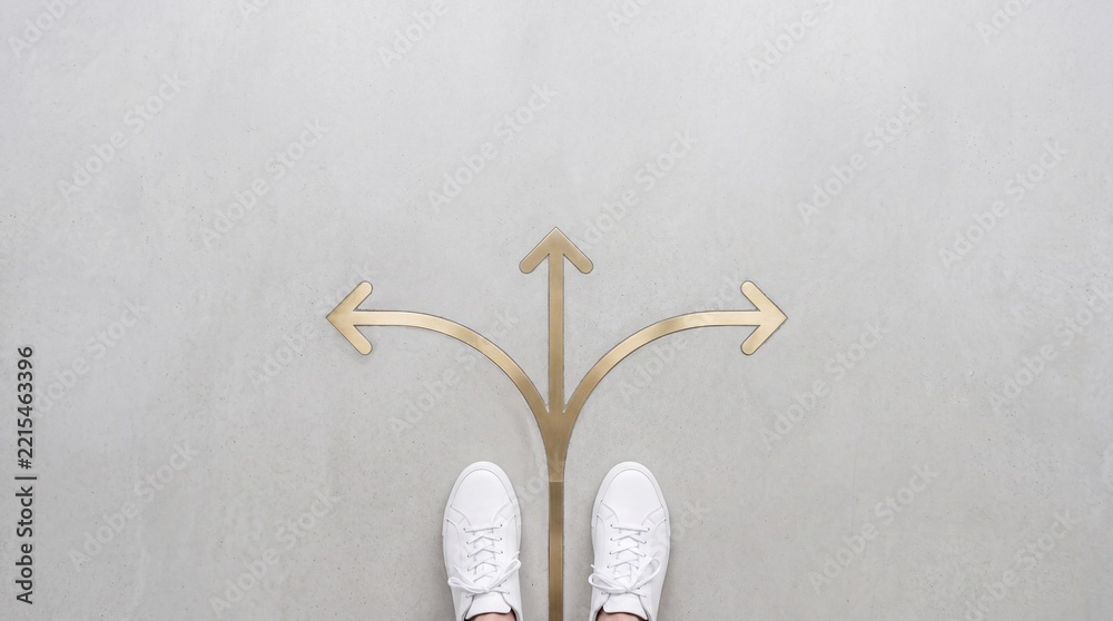 Naklejka premium Top view of a person wearing white sneakers standing at a three-way arrow crossroads on a concrete floor, representing decision making, career path options, and choosing the right direction.