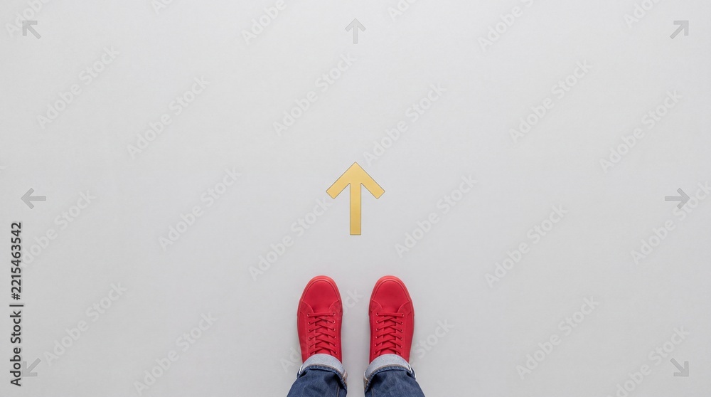 Fototapeta premium Top view of a person wearing red sneakers standing in front of a yellow forward arrow on a gray floor, representing concepts of choice, direction, decision making, and career path planning.