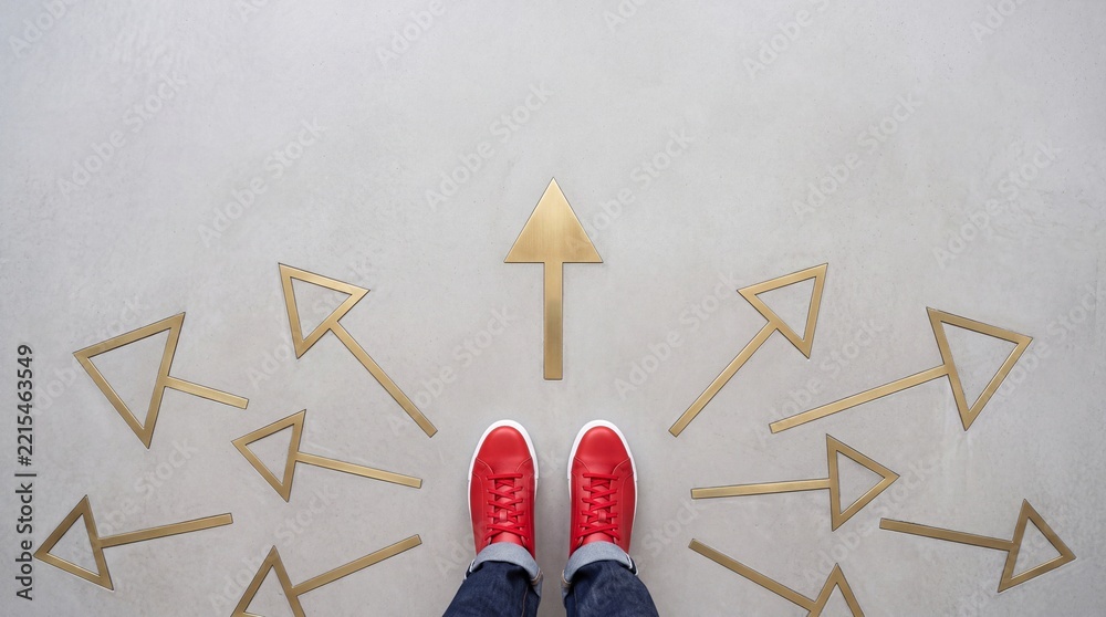 Fototapeta premium High angle top down view of a person wearing red sneakers standing on a concrete floor surrounded by multiple golden arrows pointing in different directions representing choice and career path.