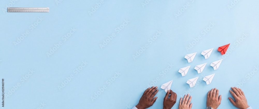 Fototapeta premium Strategic business leadership and teamwork concept featuring a red paper plane leading white ones on a wide blue background with diverse hands and a ruler for measurement and planning