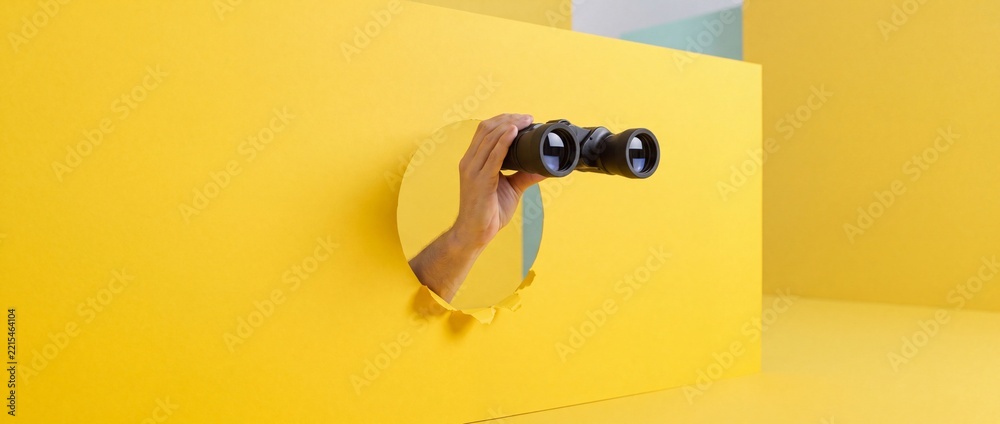 Fototapeta premium Conceptual Minimalist Image of a Person's Hand Holding Binoculars Through a Round Hole in a Bright Yellow Paper Wall Representing Discovery, Vision, Market Research, and Scouting Opportunities.