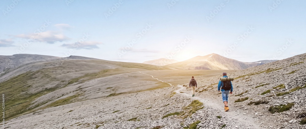 Fototapeta premium Rear view of two adventurous male hikers trekking along a winding stone path in a vast, barren mountain range under a clear sky with sunlight, ideal for travel, exploration, and outdoor lifestyle.
