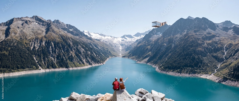 Fototapeta premium Hiker couple sitting on a cliff looking at a panoramic view of a turquoise mountain lake with a futuristic glass cabin on a ridge, wide angle landscape of the Alps adventure travel concept.