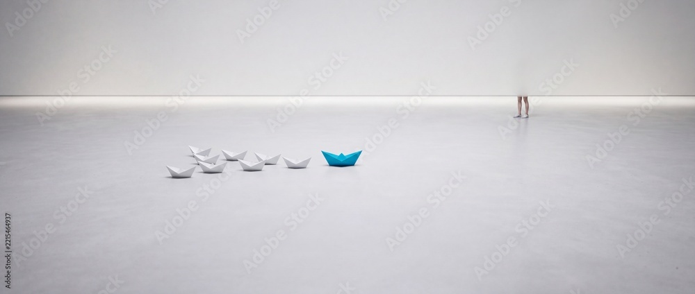 Fototapeta premium Wide conceptual shot of a blue origami paper boat leading a group of white paper boats on a minimalist white surface representing leadership, individuality, and business strategy concepts.
