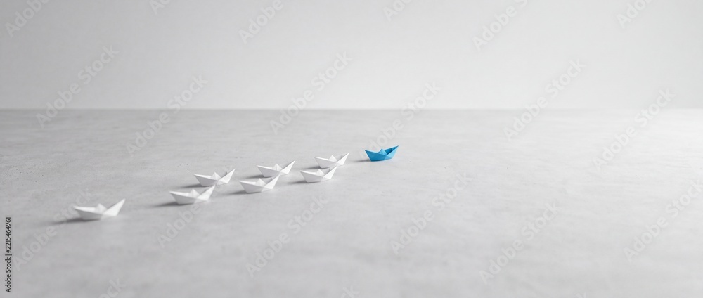 Fototapeta premium A blue origami paper boat leading a group of white paper boats on a gray textured background, representing leadership, business success, unique vision, and individuality in a minimalist wide