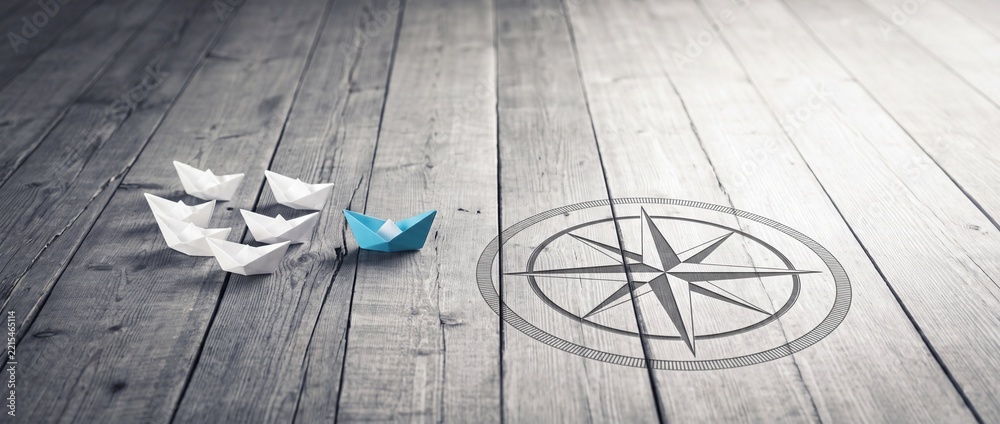 Fototapeta premium Blue Origami Paper Boat Leading a Fleet of White Boats Across a Wooden Surface Toward a Compass Symbol, Representing Leadership, Strategic Business Direction, and Vision in a Wide Panorama