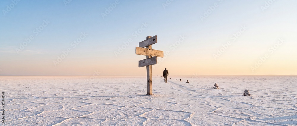 Fototapeta premium A man walking away on a vast white salt flat toward the horizon at sunset with a rustic wooden directional signpost in the foreground, symbolizing life choices, journey, and path to the future