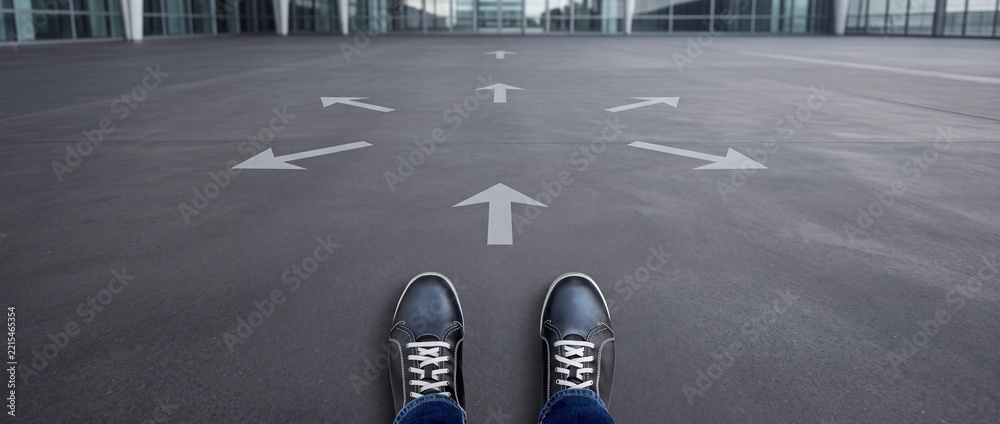 Fototapeta premium Wide angle POV of a person wearing casual shoes standing in front of several white directional arrows on asphalt, symbolizing career choices, decision making, navigation, and life path options.