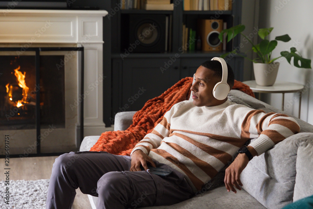 Fototapeta premium African American man reclining on light gray sofa in living room, wearing headphones