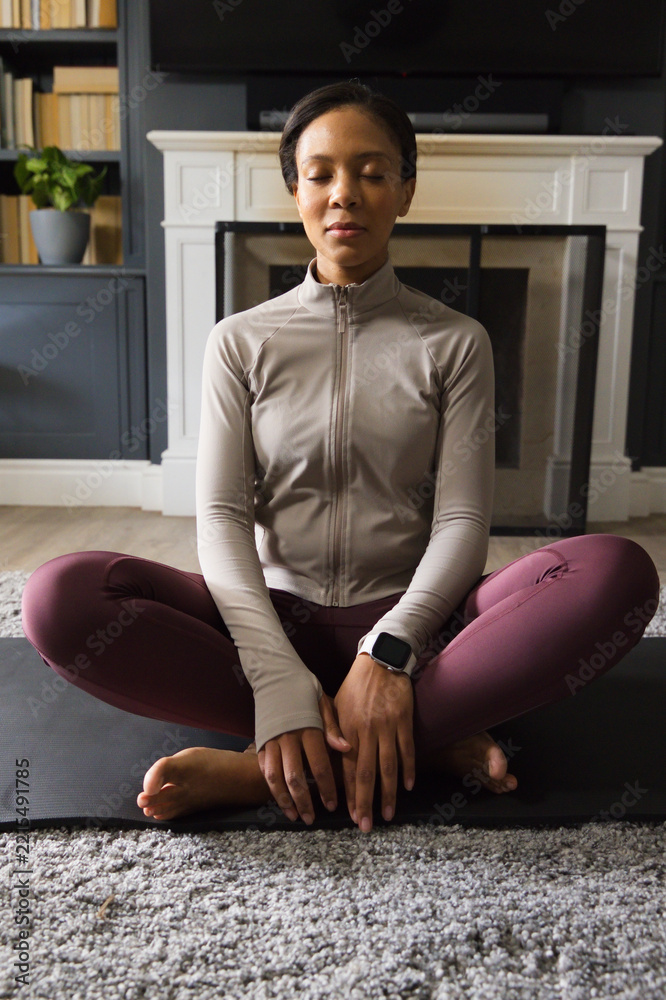Naklejka premium African American woman meditating on black yoga mat in living room, wearing leggings and smartwatch