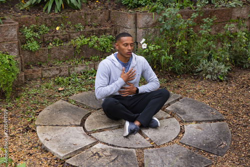 African American man sitting cross-legged meditating on stone ring in garden with hoodie smartwatch