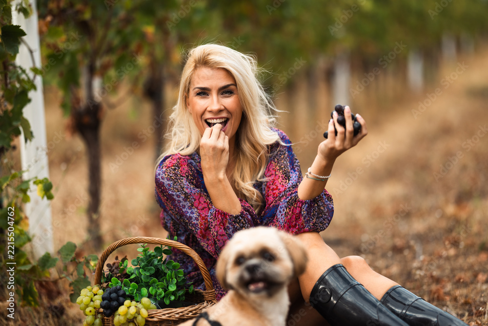 Fototapeta premium Smiling woman enjoying grapes in vineyard with dog beside her