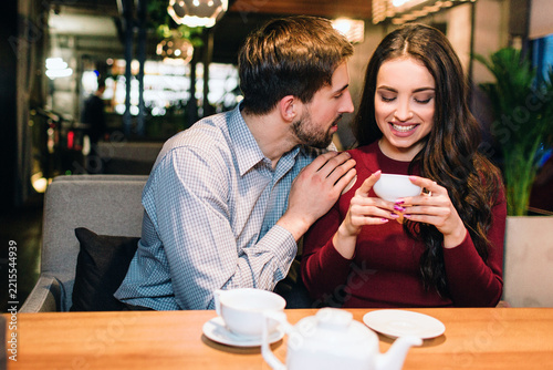 Attractive girl is sitting on the sofa in restaurant and drinking some tea while her partner has put his hand over her shoulder and looking to her. She is smiling.