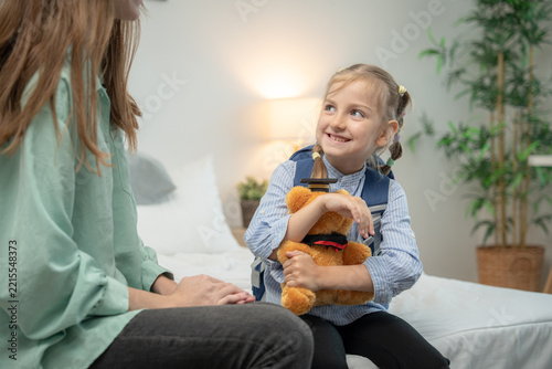 Happy little girl holding teddy bear and looking at her mother with a smile. Cheerful elementary schoolgirl wearing backpack holding graduation teddy bear and smiling at supportive mother in bedroom.