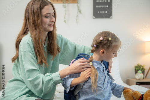 Mother supporting daughter wearing backpack while holding teddy bear on bed. Young mother smiling and supporting cute schoolgirl carrying backpack and stuffed toy bear in bedroom.
