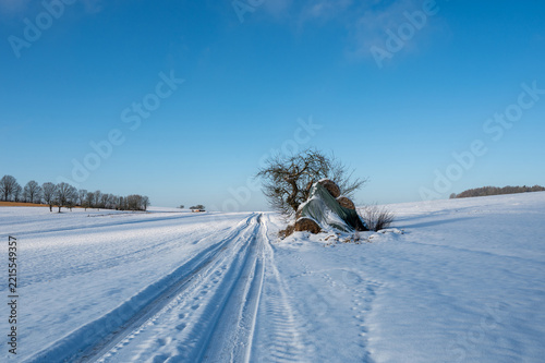 Snow-covered road in open countryside with a blue sky, a tree, and hay bales.