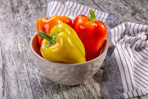 Red yellow and orange bell peppers in ceramic bowl on rustic wooden surface with striped cloth nearby vegetable display