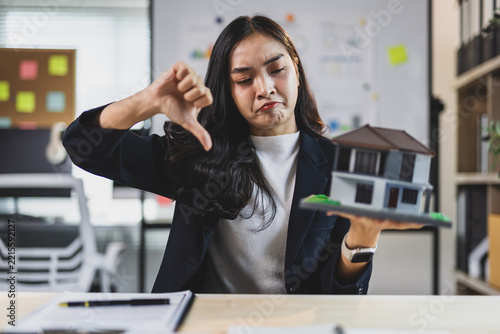 Woman agent holding a house model and giving a thumbs down gesture, expressing dissatisfaction with a property or loan deal.