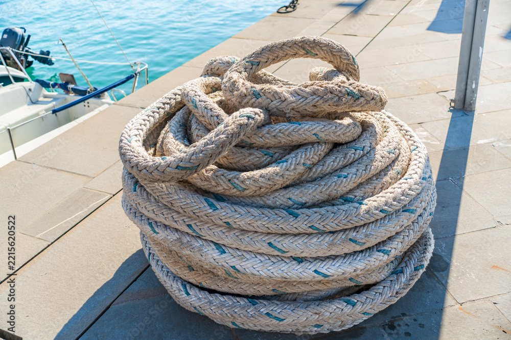 Fototapeta premium A large coil of weathered braided marine rope resting on a harbor quay beside turquoise water and a small boat