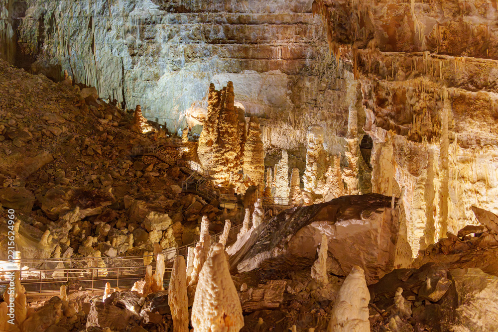 Fototapeta premium Warm light bathes ancient cave formations. Stalactites hang low, proud and tall. Stalagmites rise from earth, seeking sky. Rock walls glow with earthy hues and textures. Quiet wonder fills the dim.