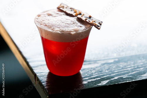 A vibrant red cocktail served in a modern glass with a thick foam layer, garnished with a piece of nut chocolate bark on the rim, resting on a dark wood table.