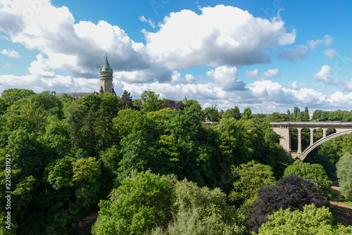 The Adolphe Bridge spanning the lush Pétrusse Valley with its iconic stone arch set against a bright summer sky in Luxembourg City.