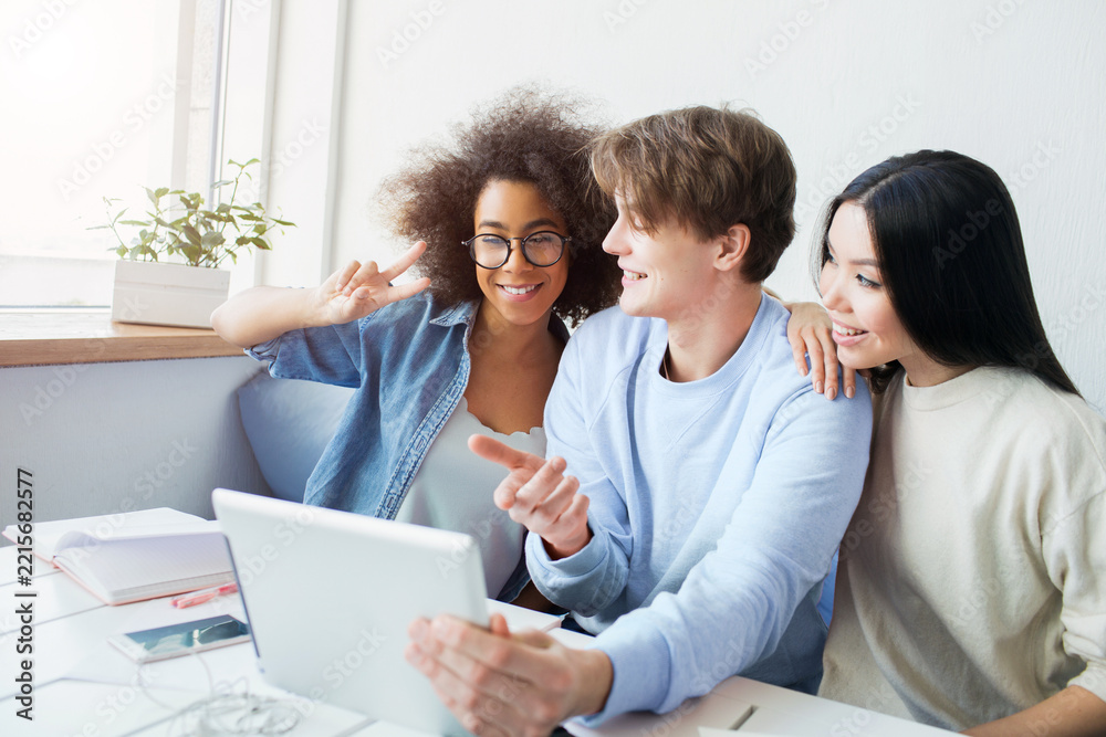 Fototapeta premium Nice picture of guy holding a tablet and girls talking with somebody using the tablet for that. Asian girl is leaning on the guy. Afro american girl is showing the piece symbol to the tablet's screen.