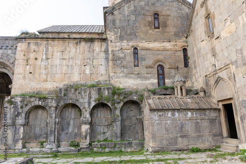 Weathered stone courtyard wall of Tatev Monastery with four blind arches and a carved tomb chapel beside tall arched windows and a doorway with weeds, Tatev, Armenia