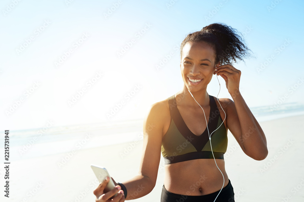 Naklejka premium Exercise, smile and woman with earphones at beach for fitness, music or workout podcast. Phone, runner and athlete with tech for marathon training, space and streaming audio or getting ready by ocean