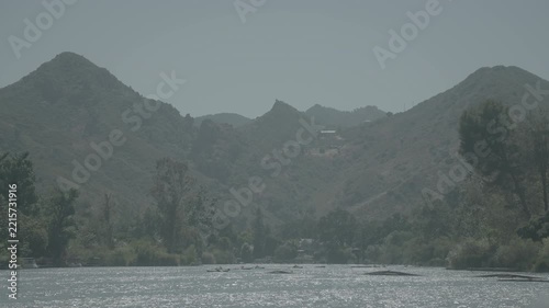 Hazy mountain peaks above green trees and a dark riverbed in soft light