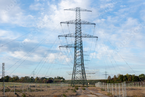 High voltage transmission tower rises at power line construction site with suspended conductors and fencing. Steel lattice structure represents grid expansion and energy engineering under cloudy sky