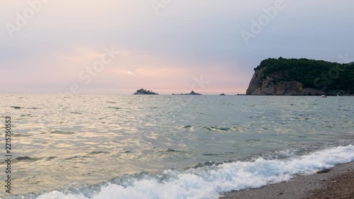 Sunset coastal wide view with gentle waves breaking on pebble beach, distant rocky headland, small islands, and calm horizon under pastel sky with soft evening glow