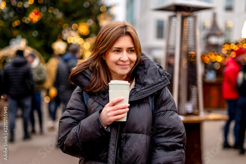 Smiling woman holding hot coffee in festive Christmas market lights during winter evening