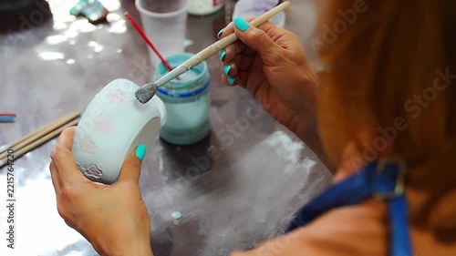 Woman applying blue glaze to handmade ceramic mug with brush. Pottery glazing process represents ceramic craft and handmade production